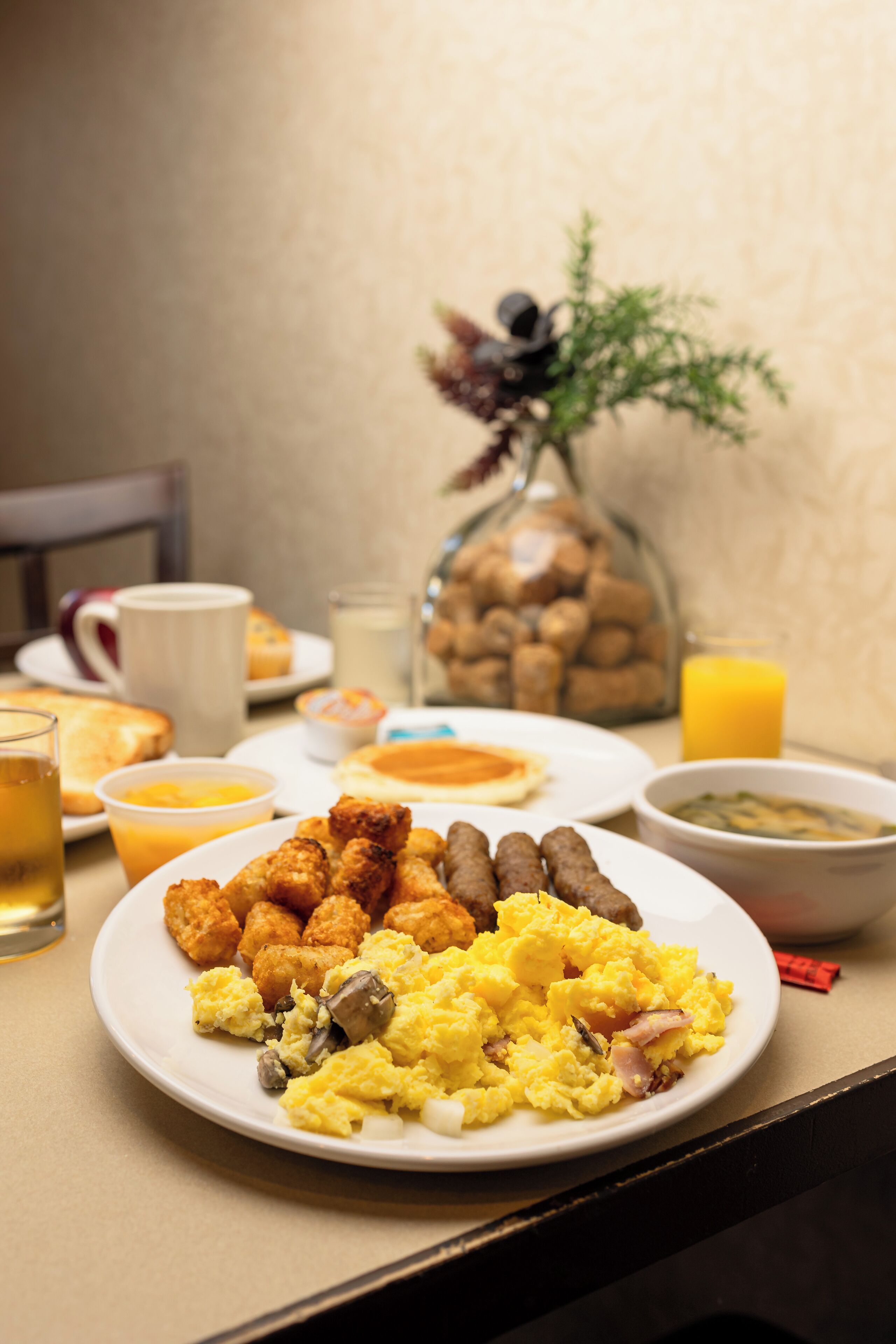 A breakfast table with plates of scrambled eggs, sausages, and tater tots, along with a glass of orange juice and a coffee mug.