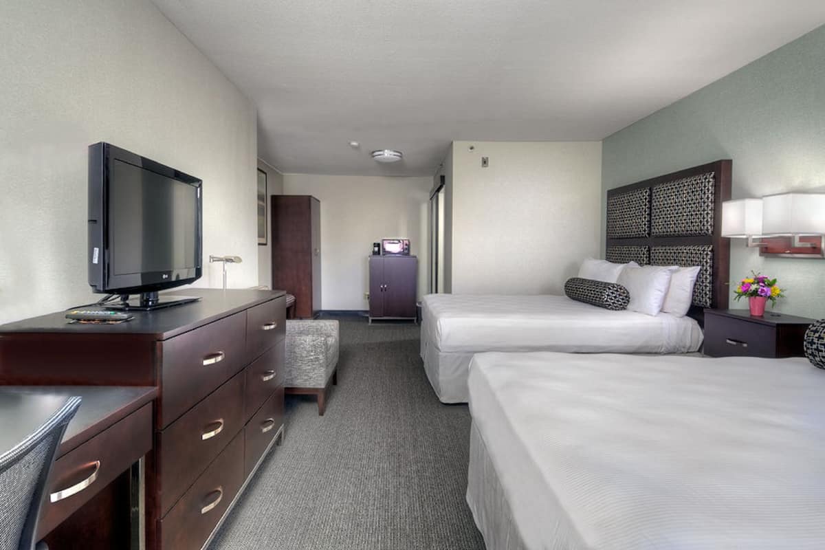 A hotel room with two beds, a desk with a television, a dresser, a small cabinet, and a nightstand with a lamp.