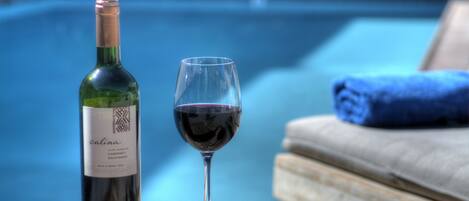A bottle of wine and a glass of red wine on a wooden table by a poolside.