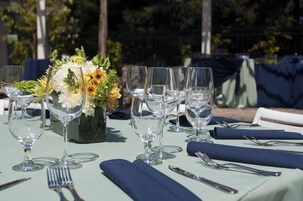 Una mesa dispuesta para un evento al aire libre formal, con copas transparentes, servilletas azules y un centro de mesa floral.