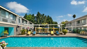 A swimming pool with lounge chairs and umbrellas, surrounded by a building with multiple windows and a covered seating area. - Maple Tree Inn (Sunnyvale)