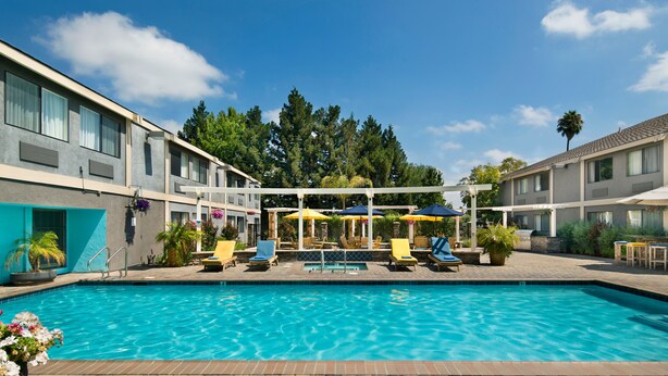 A swimming pool with lounge chairs and umbrellas, surrounded by a building with multiple windows and a covered seating area.