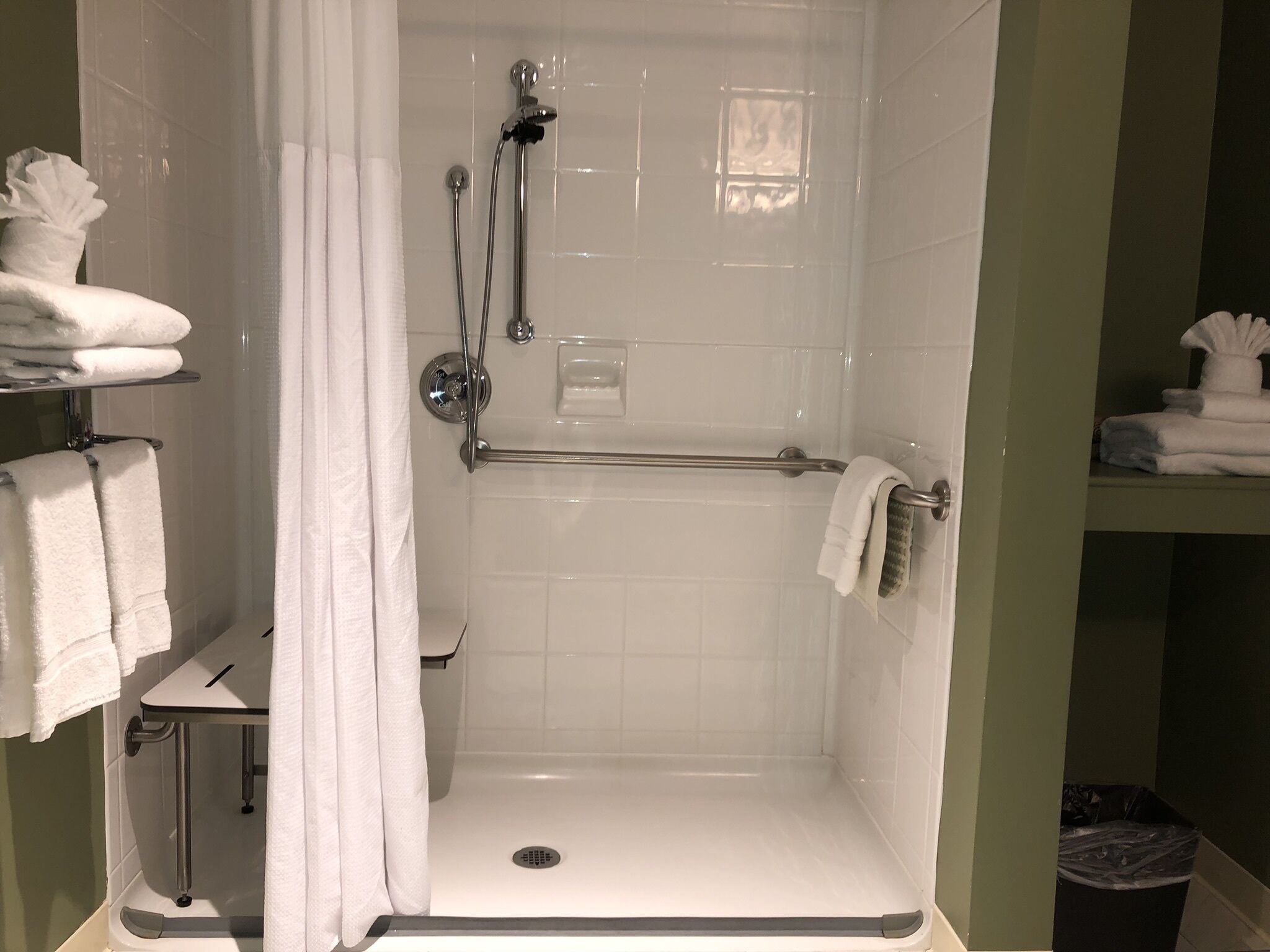 Ducha de azulejos blancos con cabezal de ducha de mano, barra de apoyo y toallero con toallas dobladas.
