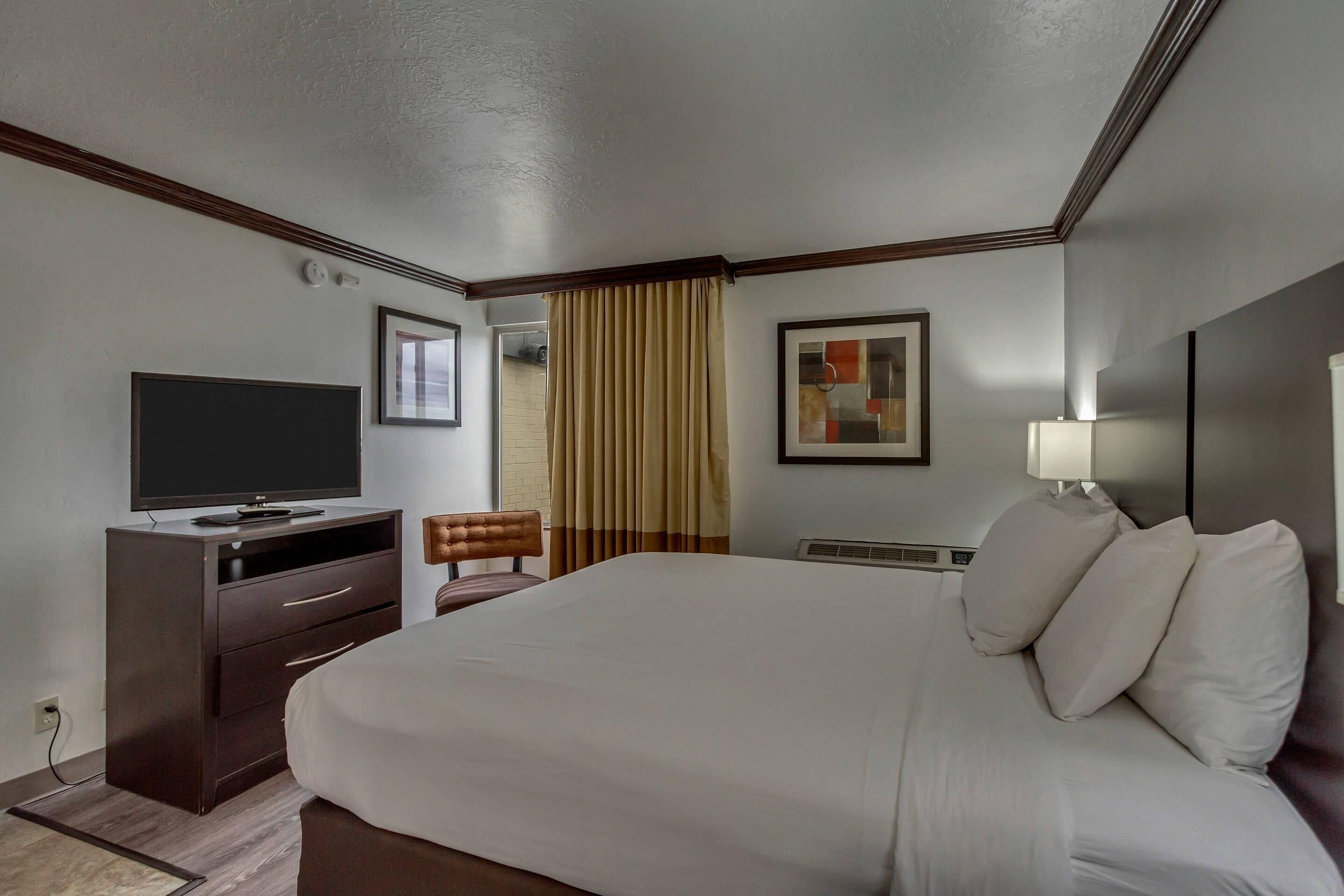 A hotel room with a large bed, a TV on a dresser, a chair, a window with curtains, and a framed picture on the wall.