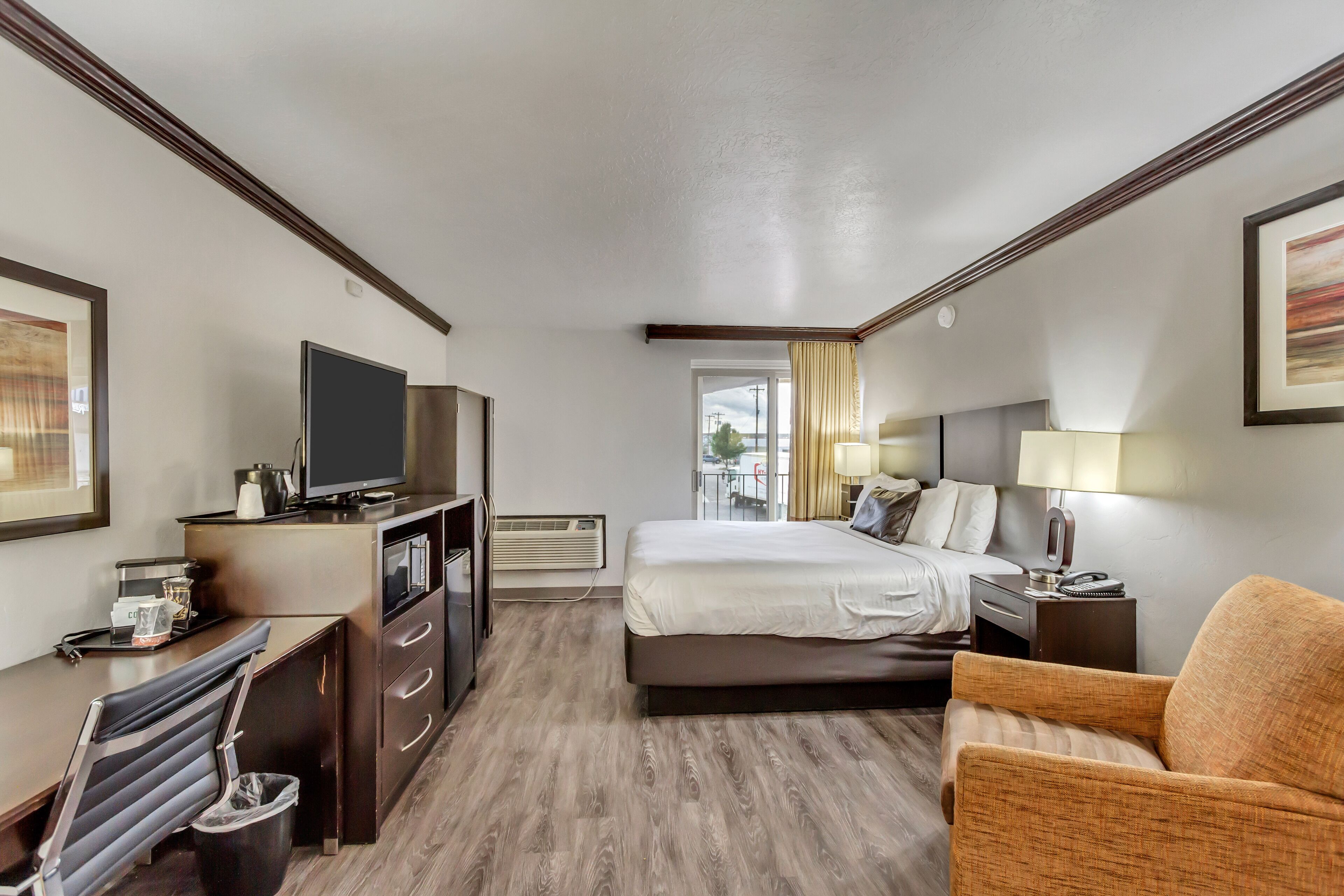 A hotel room with a bed, a desk with a TV, a chair, a bedside table with a lamp, and a window with curtains.