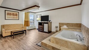 Habitación de hotel con suelo de madera, sofá, televisor, refrigerador pequeño, baño con bañera y ventana con cortinas.