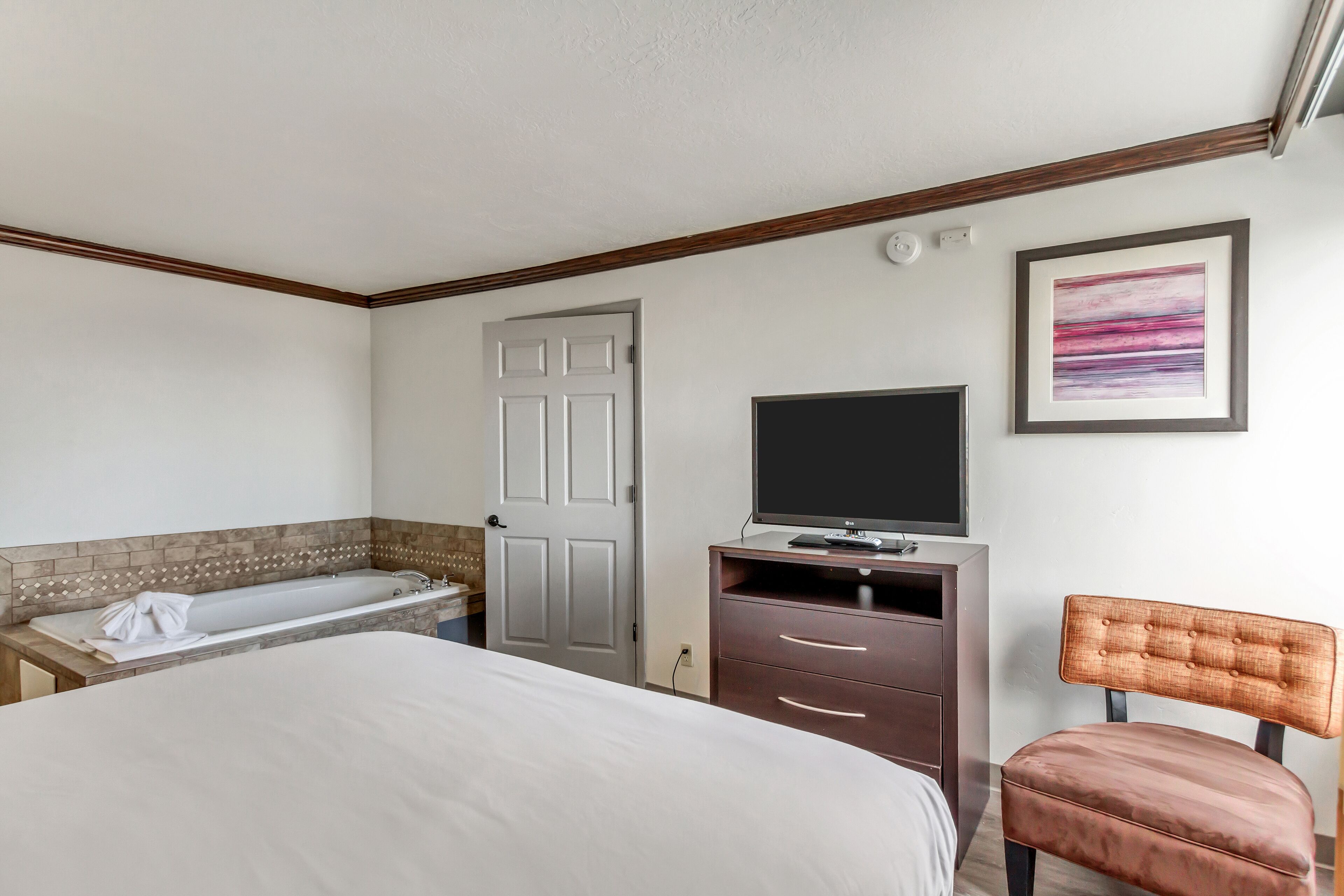 Une chambre d’hôtel comprenant un lit, un bureau avec un téléviseur à écran plat, une chaise, une porte et un tableau encadré au mur.