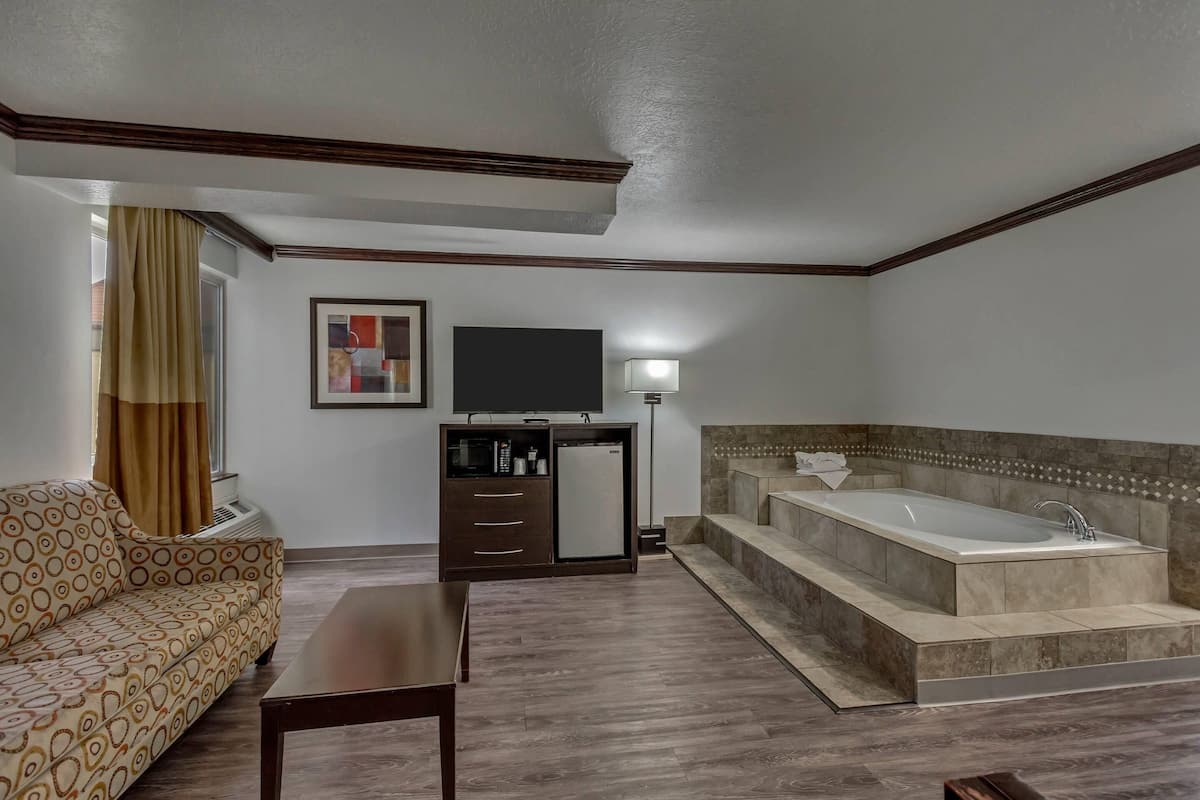 A hotel room with a sofa, a flat-screen TV, a minibar, a bathtub, and a lamp.