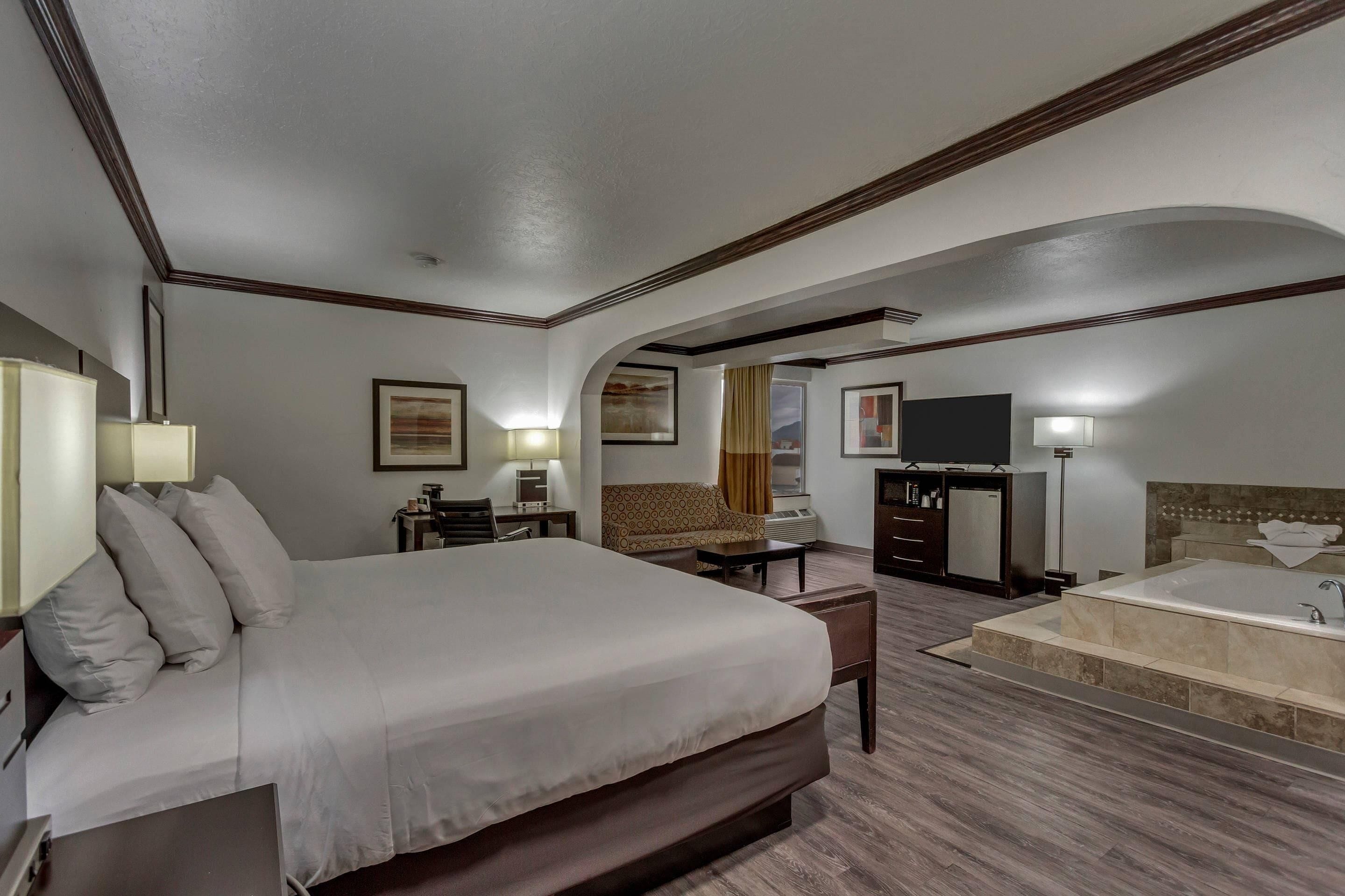 Une chambre d’hôtel dotée d’un grand lit, d’un coin salon avec un canapé et des fauteuils, d’un bureau avec une chaise, d’une coiffeuse avec un miroir, d’une grande baignoire et d’une fenêtre avec des rideaux.