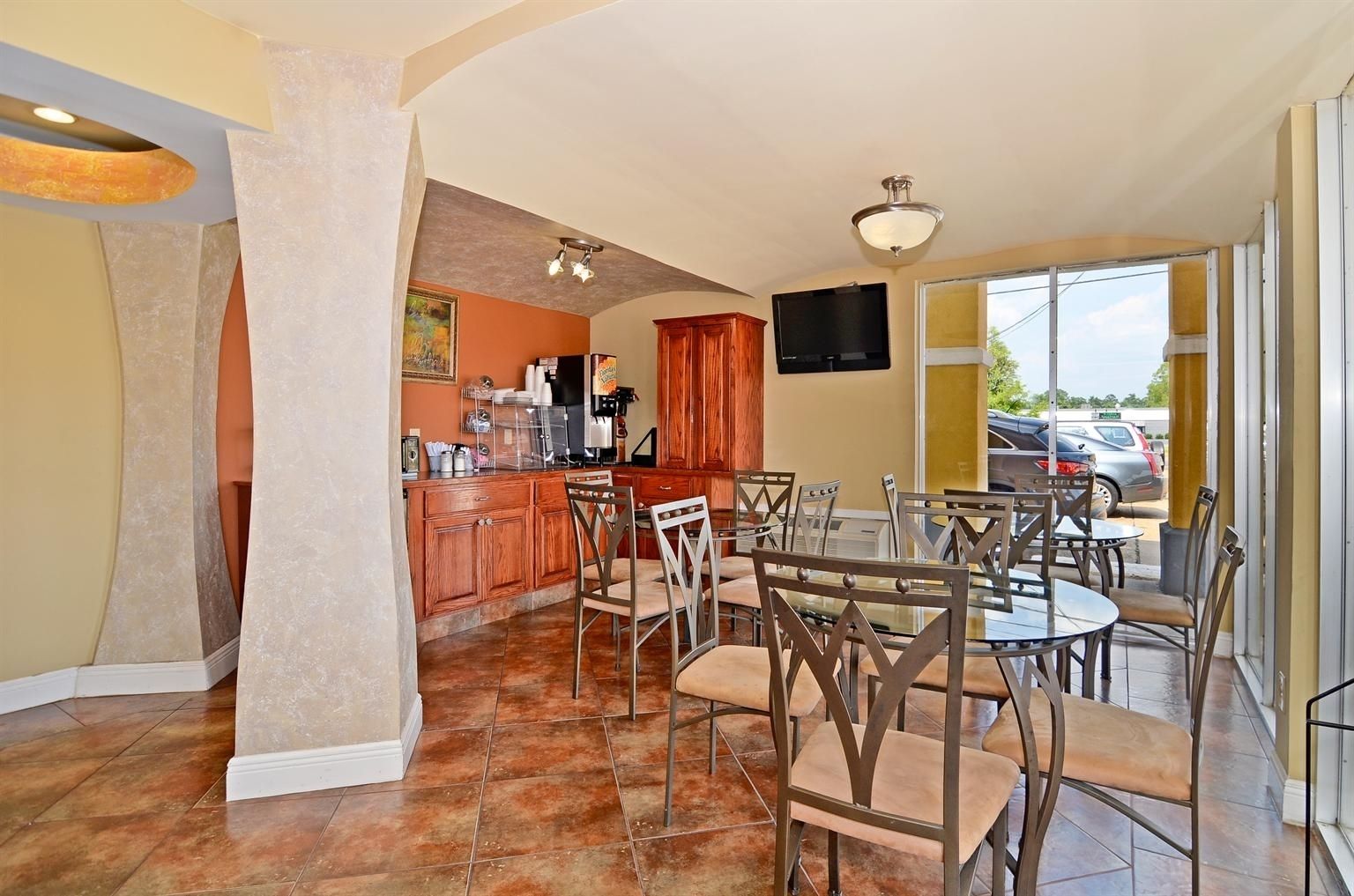 A dining area with a kitchen in the background, a TV mounted on the wall, and a view of parked cars outside.