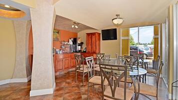 A dining area with a kitchen in the background, a TV mounted on the wall, and a view of parked cars outside.