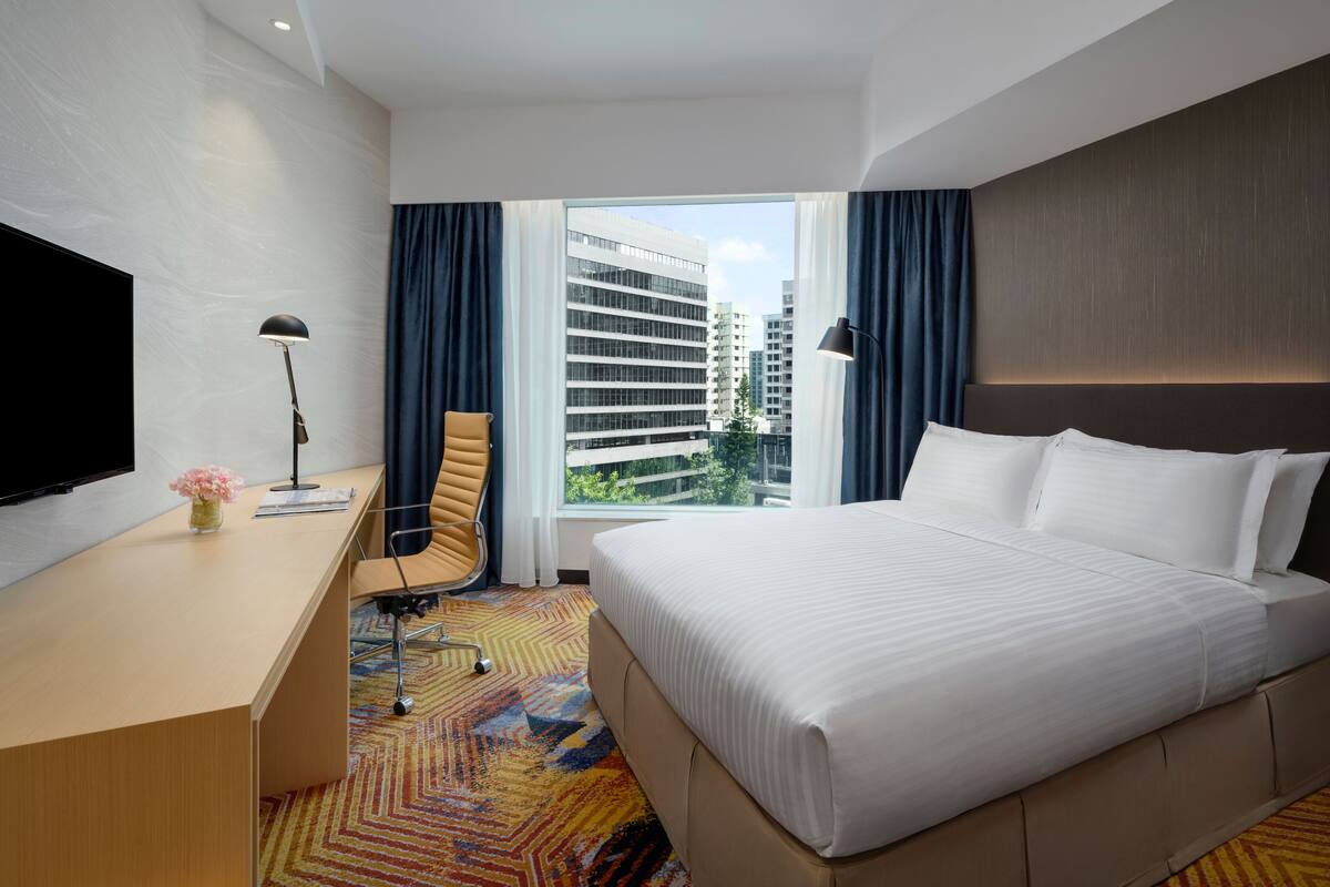 A hotel room with a large bed, a desk, a chair, a television, and a window with a view of buildings.