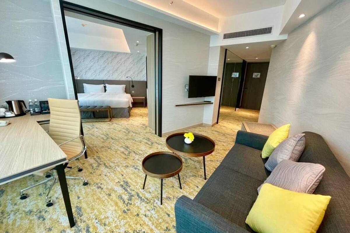 A modern hotel room with a bed, a sofa, a dining table, and a TV.
