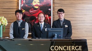 Tres hombres con traje en un mostrador de recepción con una computadora Lenovo, un cartel de fondo y un letrero que dice 'CONCIERGE'.