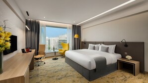 Un hotel moderno con una cama grande, un escritorio con una silla, una mesita con una lámpara y vistas de la ciudad a través de amplias ventanas.
