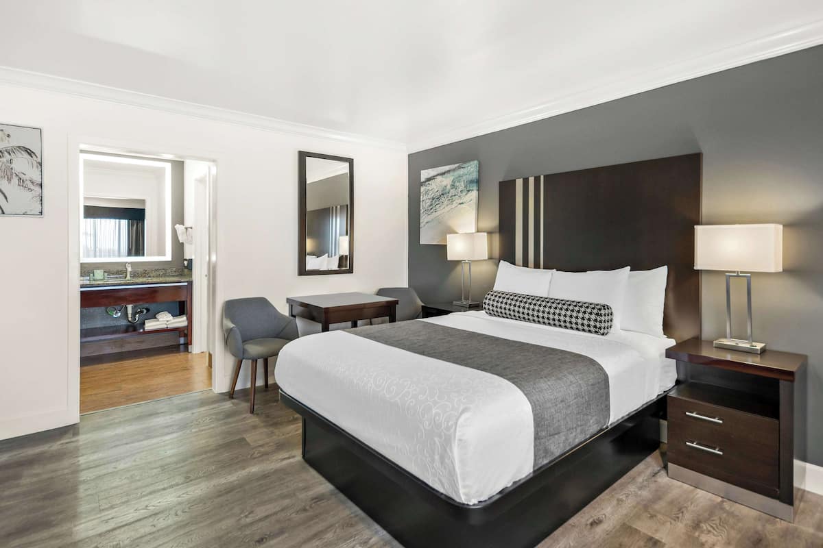 A modern hotel room with a large bed, bedside tables, a desk, and a bathroom visible through an open door.