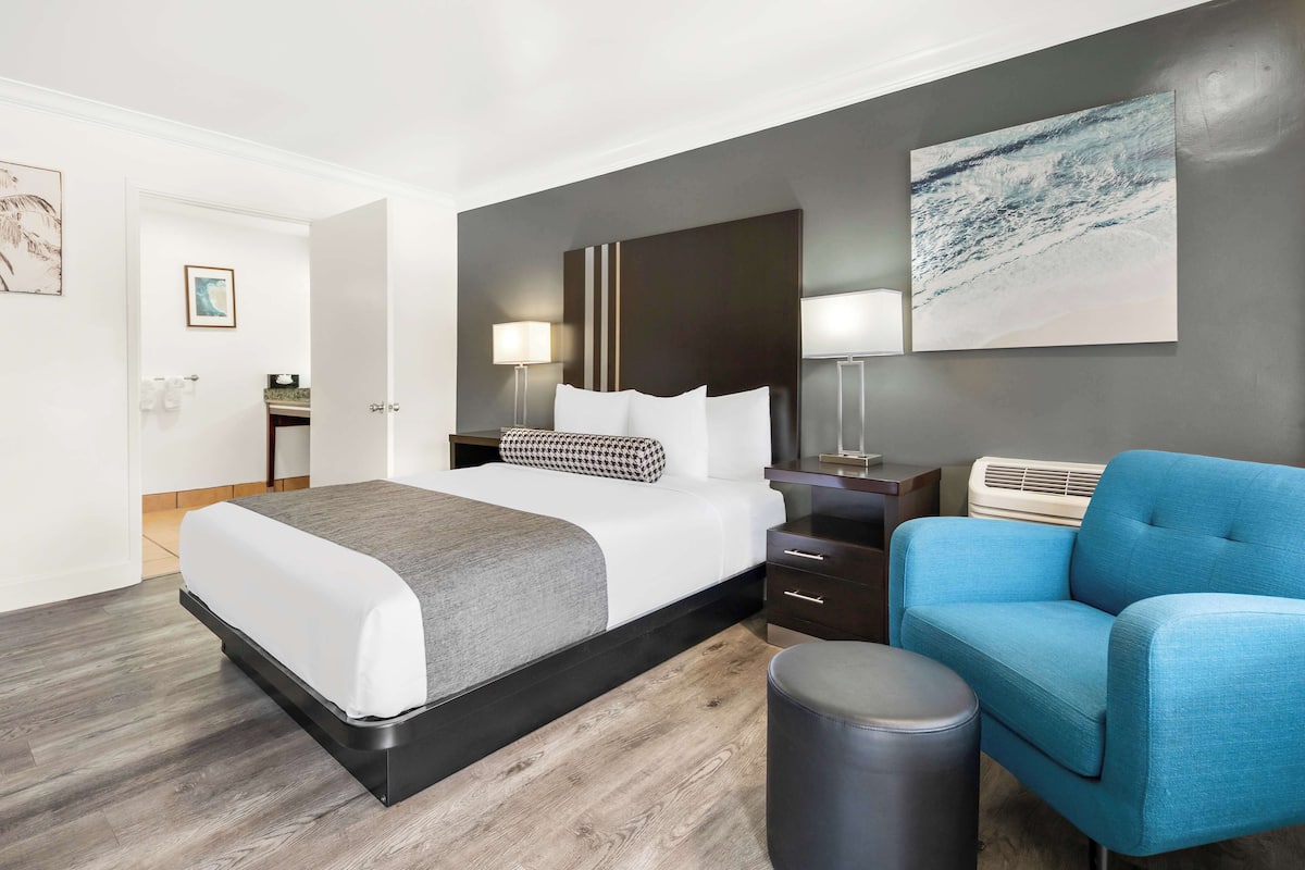 A modern hotel room with a large bed, bedside tables, a blue armchair, and a wall-mounted air conditioner.