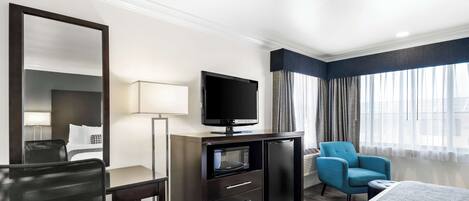 A hotel room with a bed, a desk with a television, a chair, and a minibar.