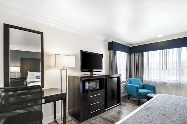 A hotel room with a bed, a desk with a television, a chair, and a minibar.