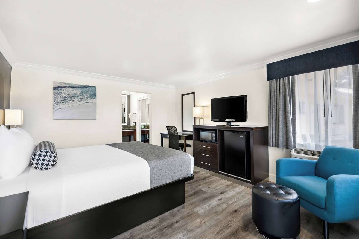 A hotel room with a bed, a desk, a television, a blue armchair, and a window with curtains.