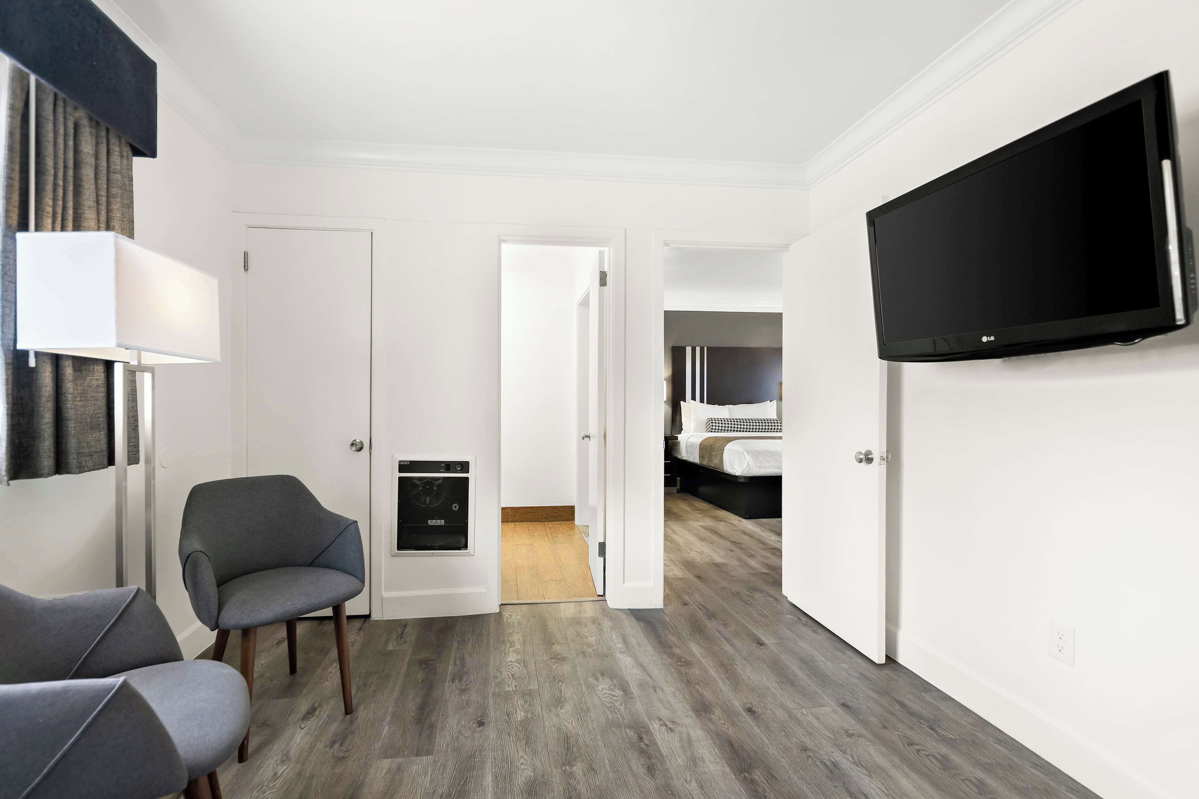 Quarto de hotel com televisão de tela plana, uma poltrona cinza, piso de madeira e uma porta que dá para outro cômodo.