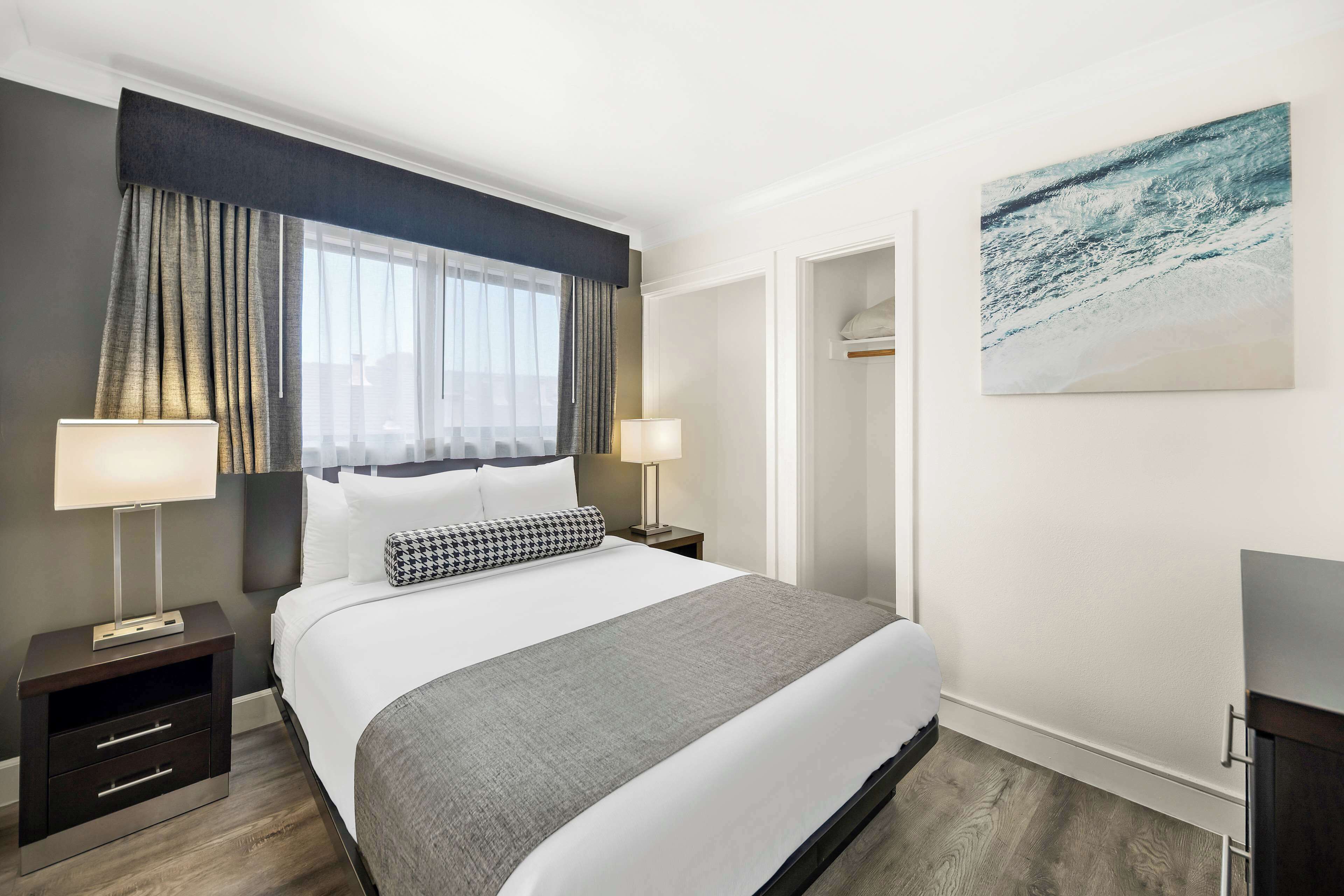 A modern hotel room with a large bed, two bedside tables with lamps, a grey throw blanket, and a painting on the wall.