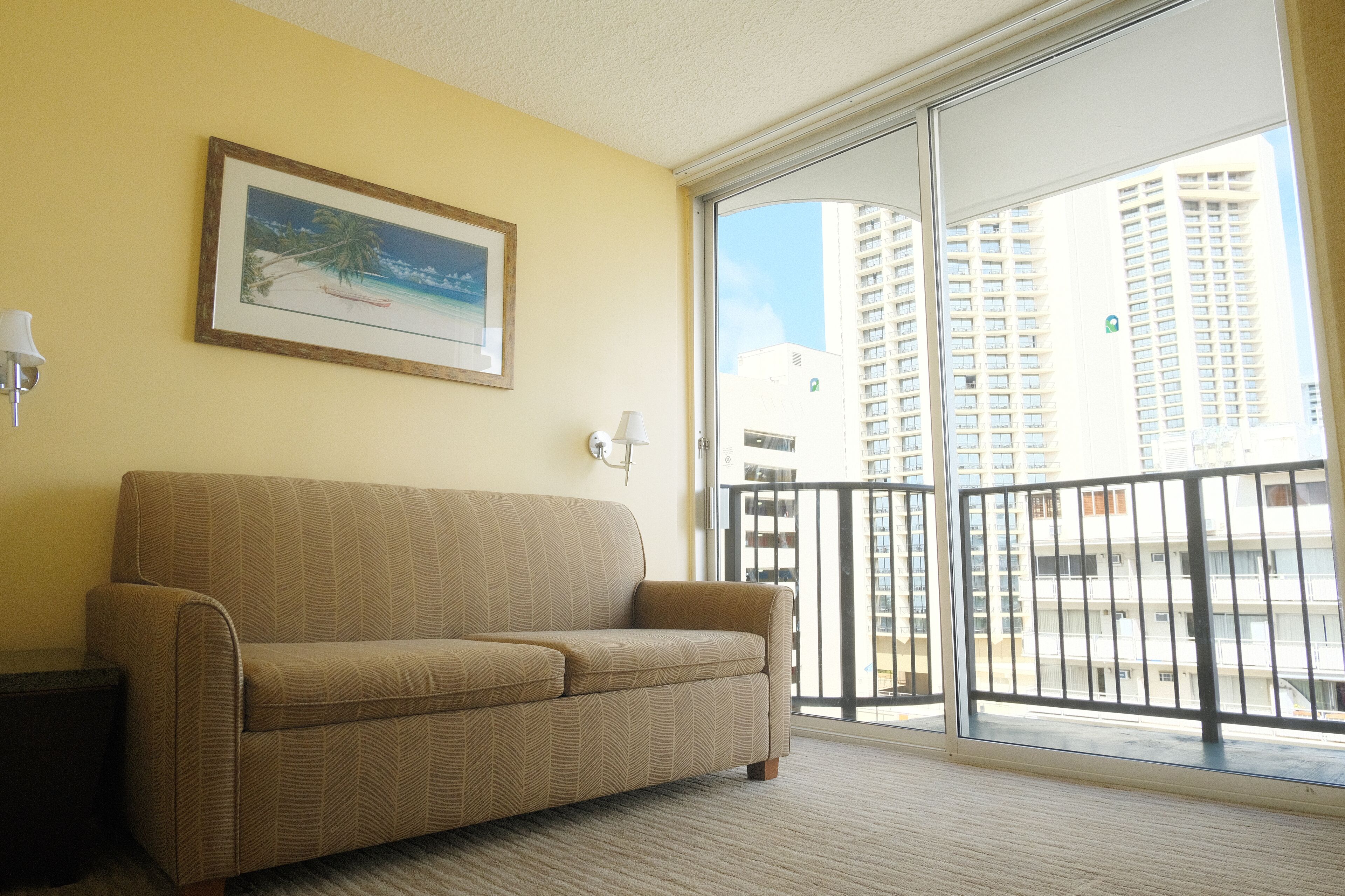 Uma sala de estar com um sofá bege, um quadro com uma cena de praia, uma mesinha com uma luminária e vista para prédios altos através de uma porta de vidro.