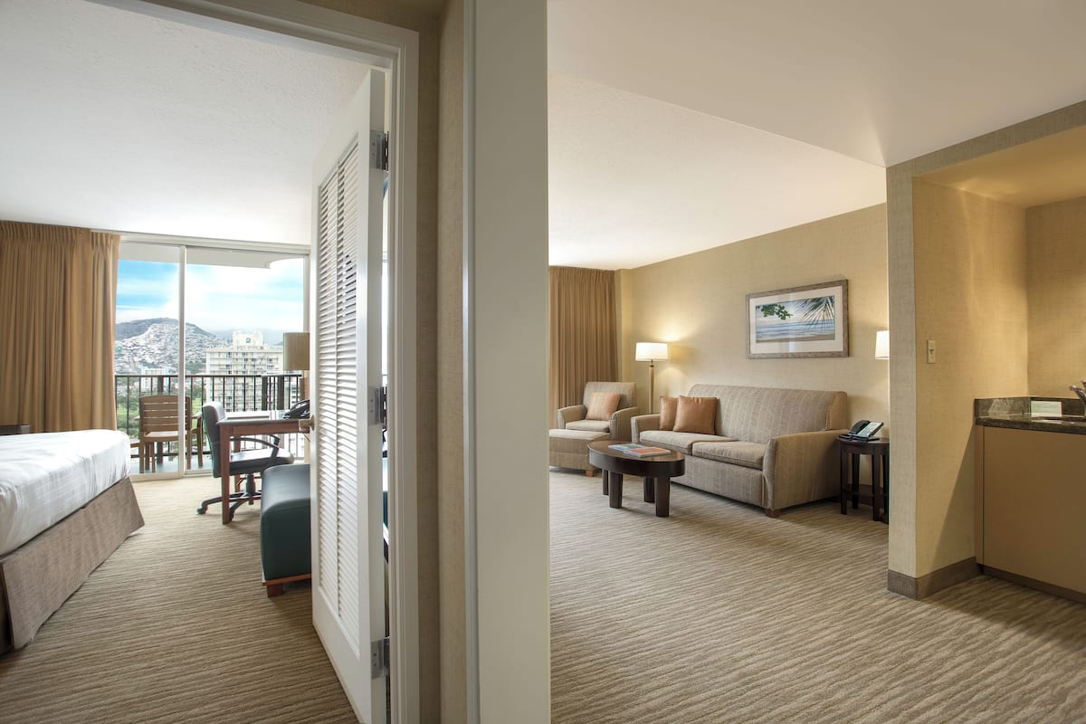 Ein Hotelzimmer mit Balkon, einem Bett, einem Schreibtisch, einem Sessel, einer Couch, einem Couchtisch und einer kleinen Küchenzeile.