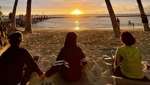 A beach scene with palm trees, a sunset, and people sitting on mats. - Waikiki Resort Hotel (Honolulu)