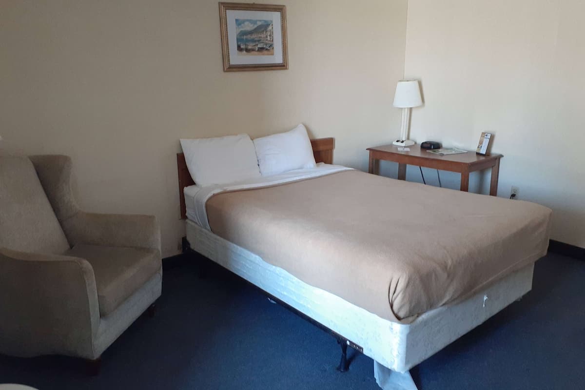 A hotel room with a bed, a chair, a bedside table with a lamp, and a framed picture on the wall.