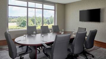 A conference room with a large table, six chairs, a flat-screen TV, and a view of greenery through large windows.
