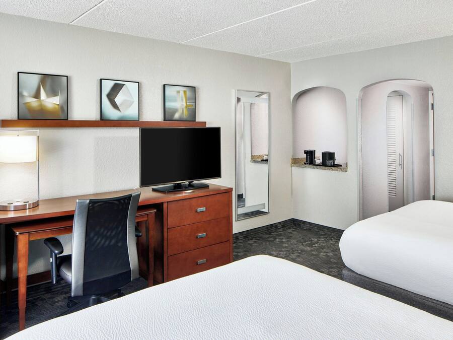 A hotel room with a desk, chair, TV, two beds, a mirror, and artwork on the wall.