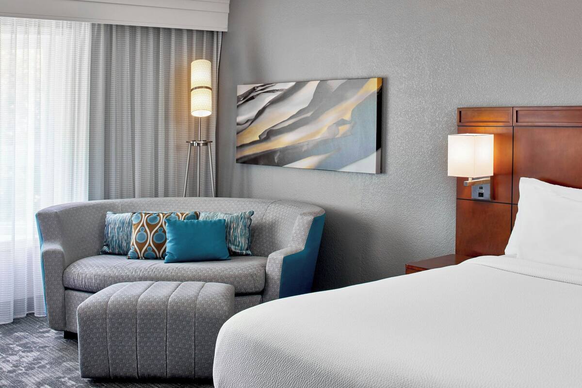 Habitación de hotel con sofá gris, un otoman a juego, cama con ropa blanca, un cuadro abstracto colgado en la pared, una lámpara de pie y una mesita de noche con lámpara.