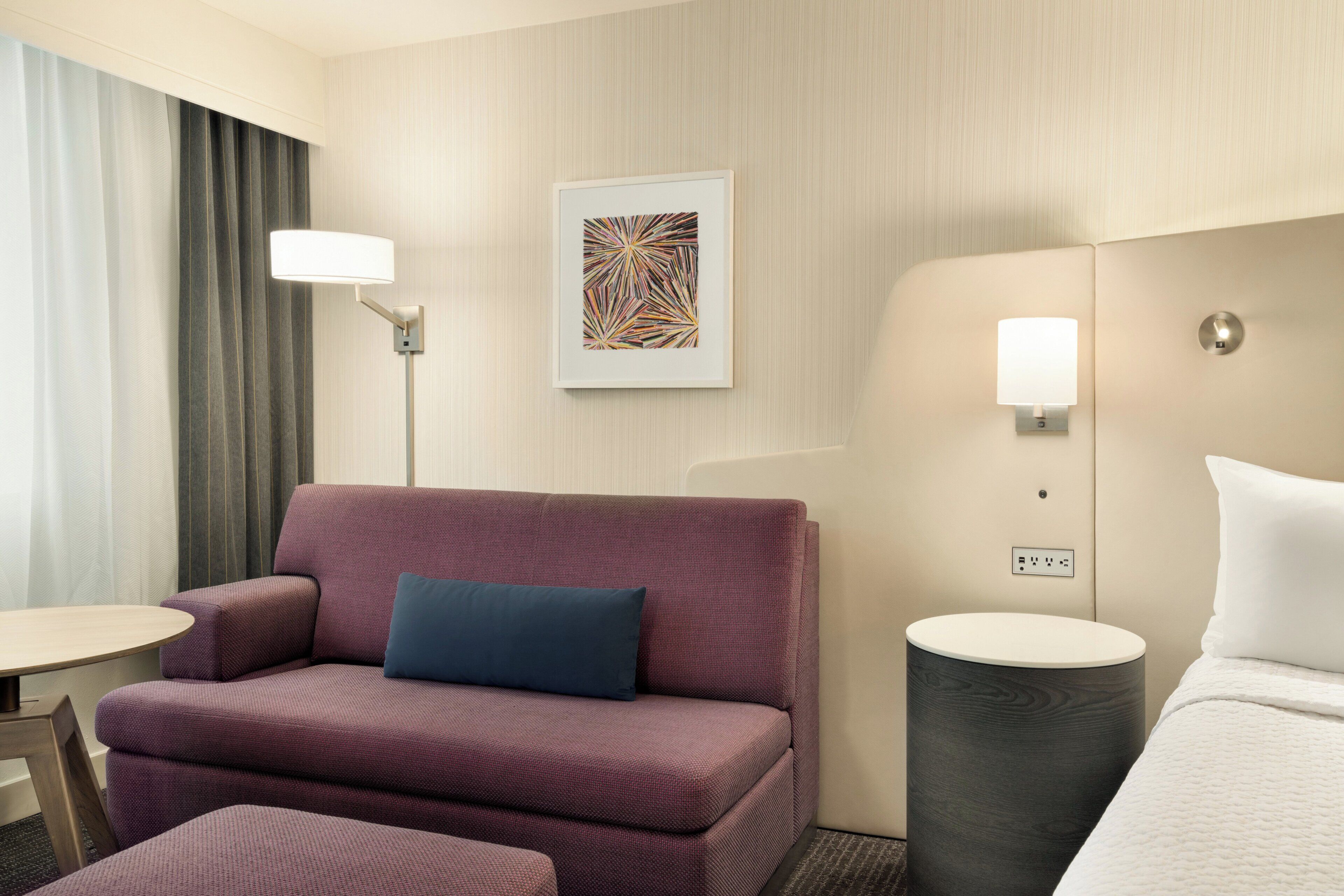 A hotel room with a sofa, a small table, a lamp, a painting, and a bedside table with a lamp.