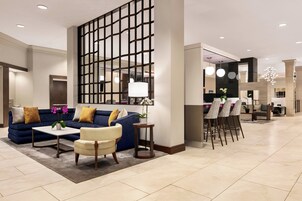 Un vestíbulo de hotel moderno con un sofá azul, un sillón blanco, una mesita y un separador decorativo.