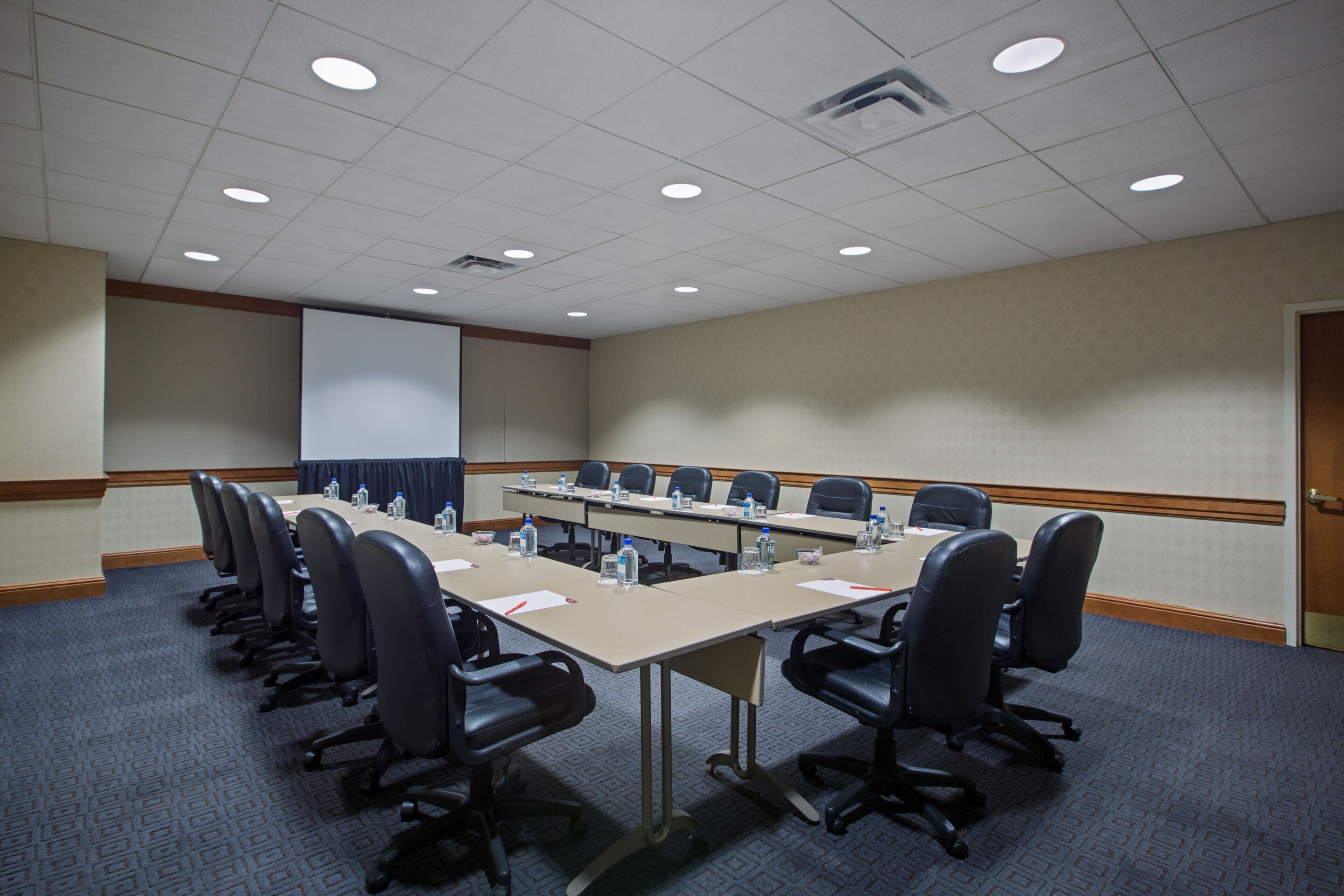 A conference room with a long table, multiple chairs, a projector screen, and a carpeted floor.
