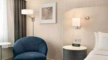 A hotel room with a blue armchair, a bedside table with a lamp, a framed abstract artwork, and a bed with white bedding.