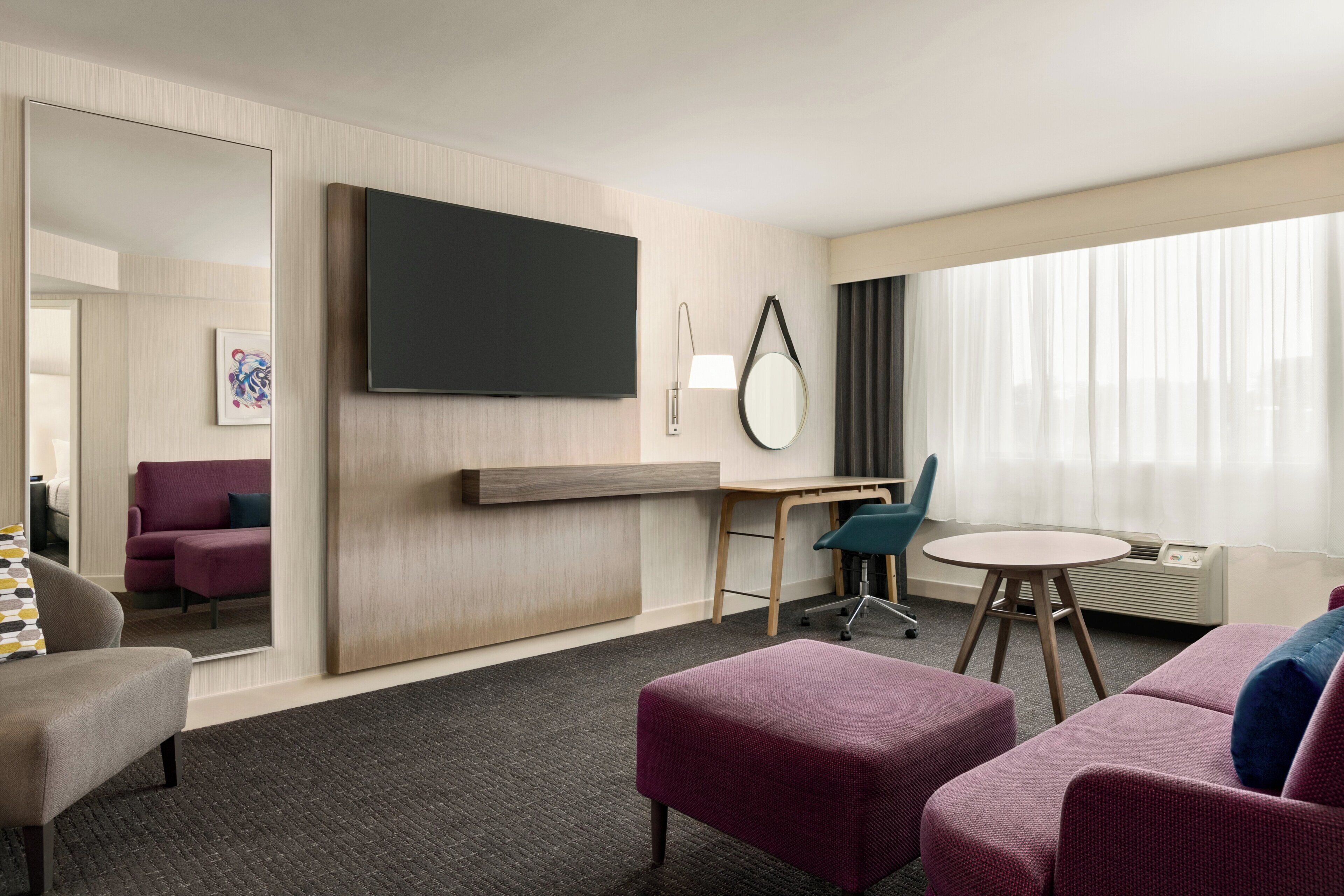A hotel room with a flat-screen TV, a desk with a chair, a round table, a sofa, and a large mirror.