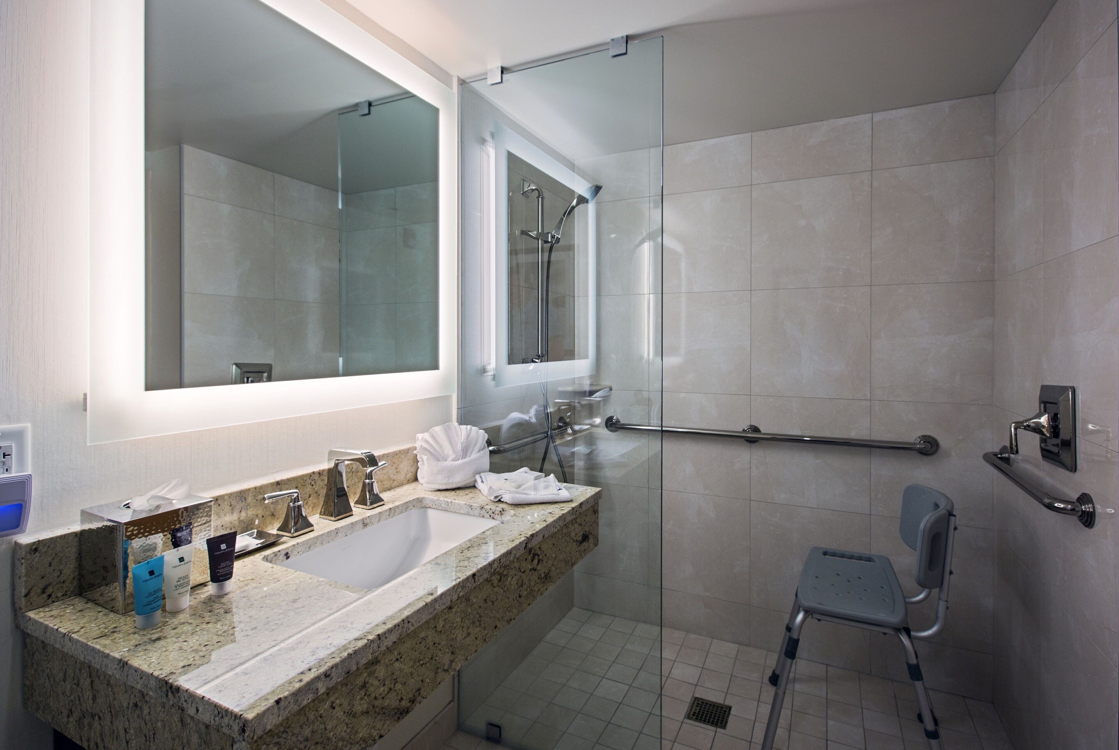 A modern bathroom with a granite countertop, a glass shower enclosure, a grab bar, and a wheelchair-accessible chair.
