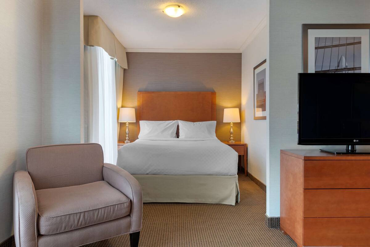 Ein Hotelzimmer mit einem Bett, zwei Nachttischlampen, einem Fernseher auf einer Holzkommode und einem Sessel.