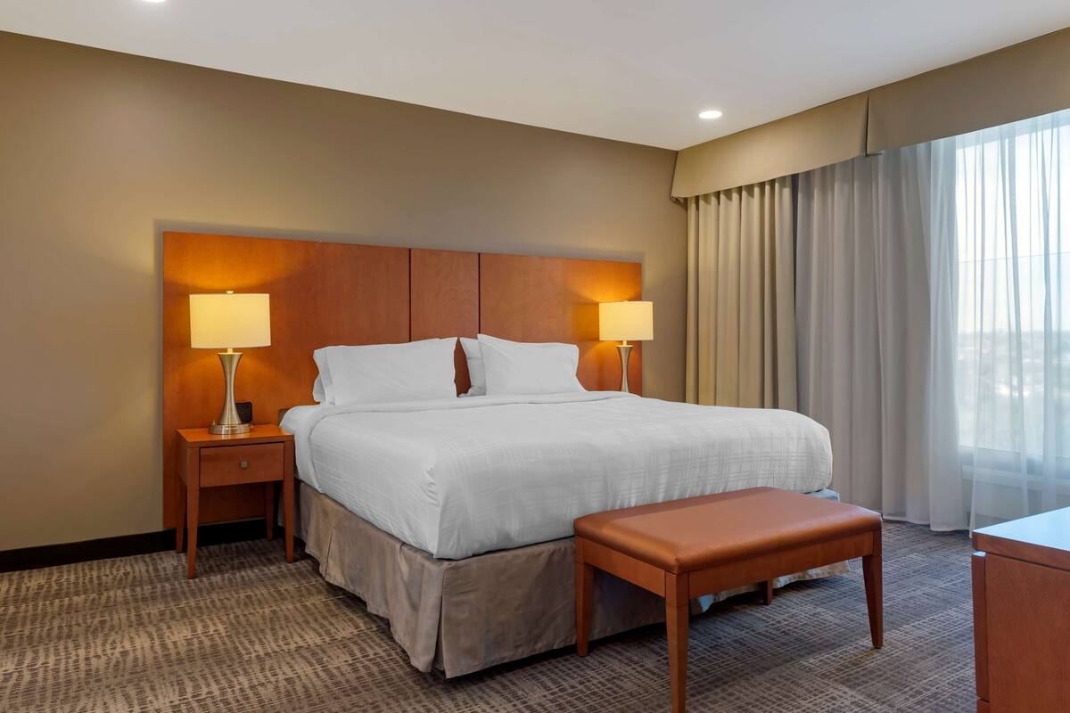 Ein Hotelzimmer mit einem großen Bett, zwei Nachttischlampen, einem Holzkopfstück, einer kleinen Holzbank und einem großen Fenster mit Vorhängen.