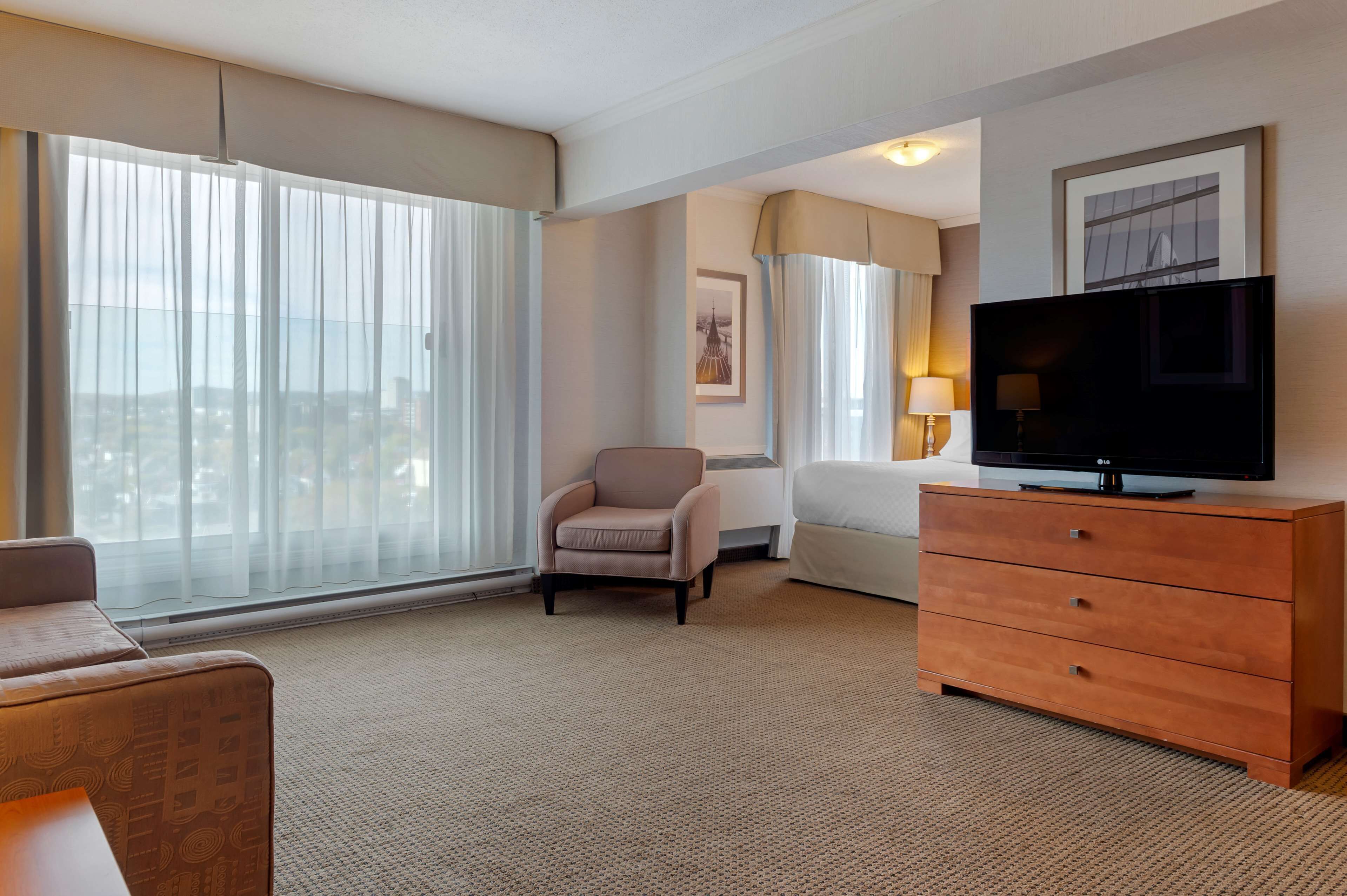 Una habitación de hotel con un ventanal grande, una televisión, un sofá, una silla y un aparador de madera.