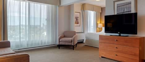 Una habitación de hotel con un ventanal grande, una televisión, un sofá, una silla y un aparador de madera.