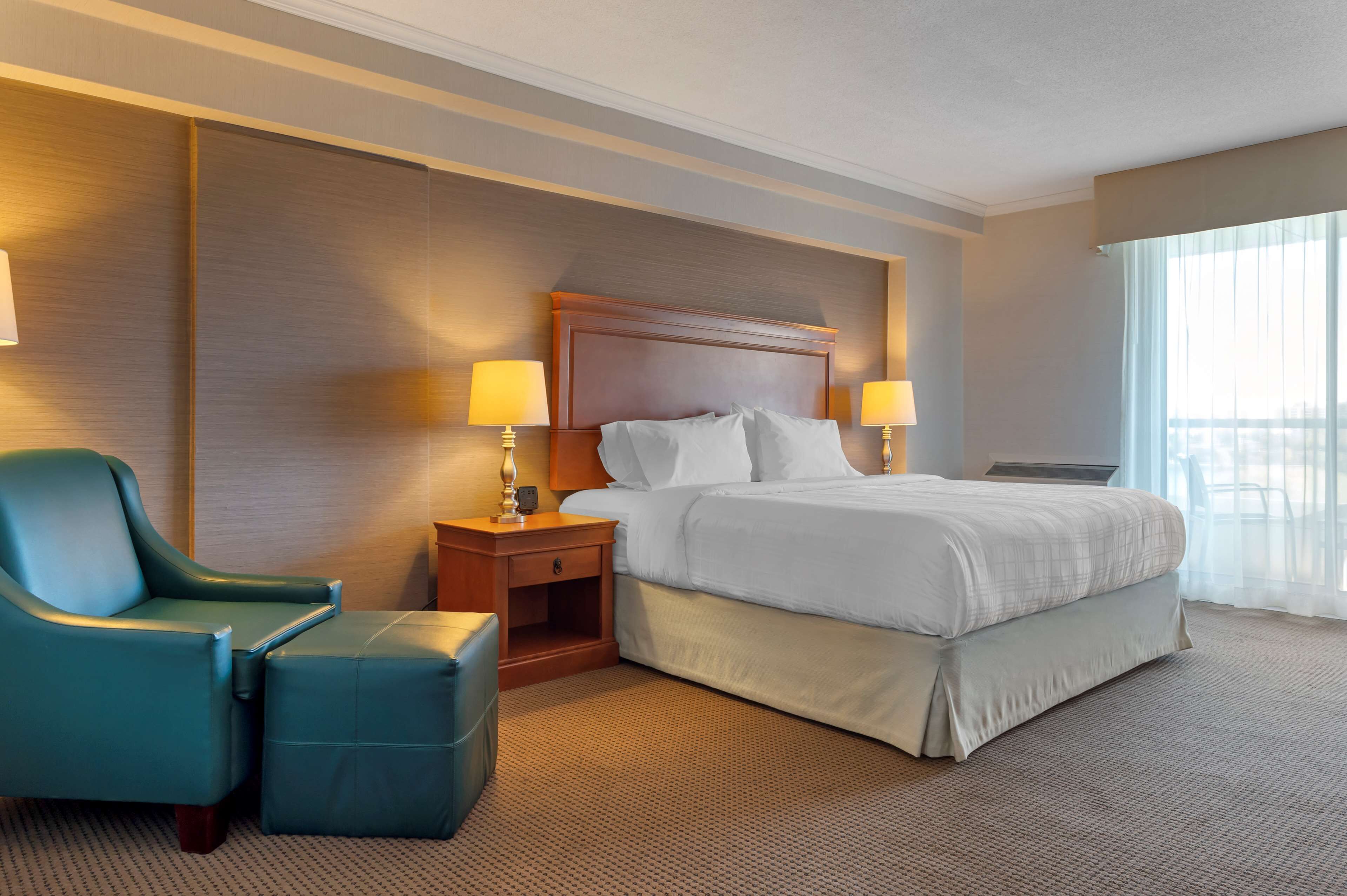 A hotel room with a large bed, a nightstand with lamps, a chair, and a view of the outdoors through the window.