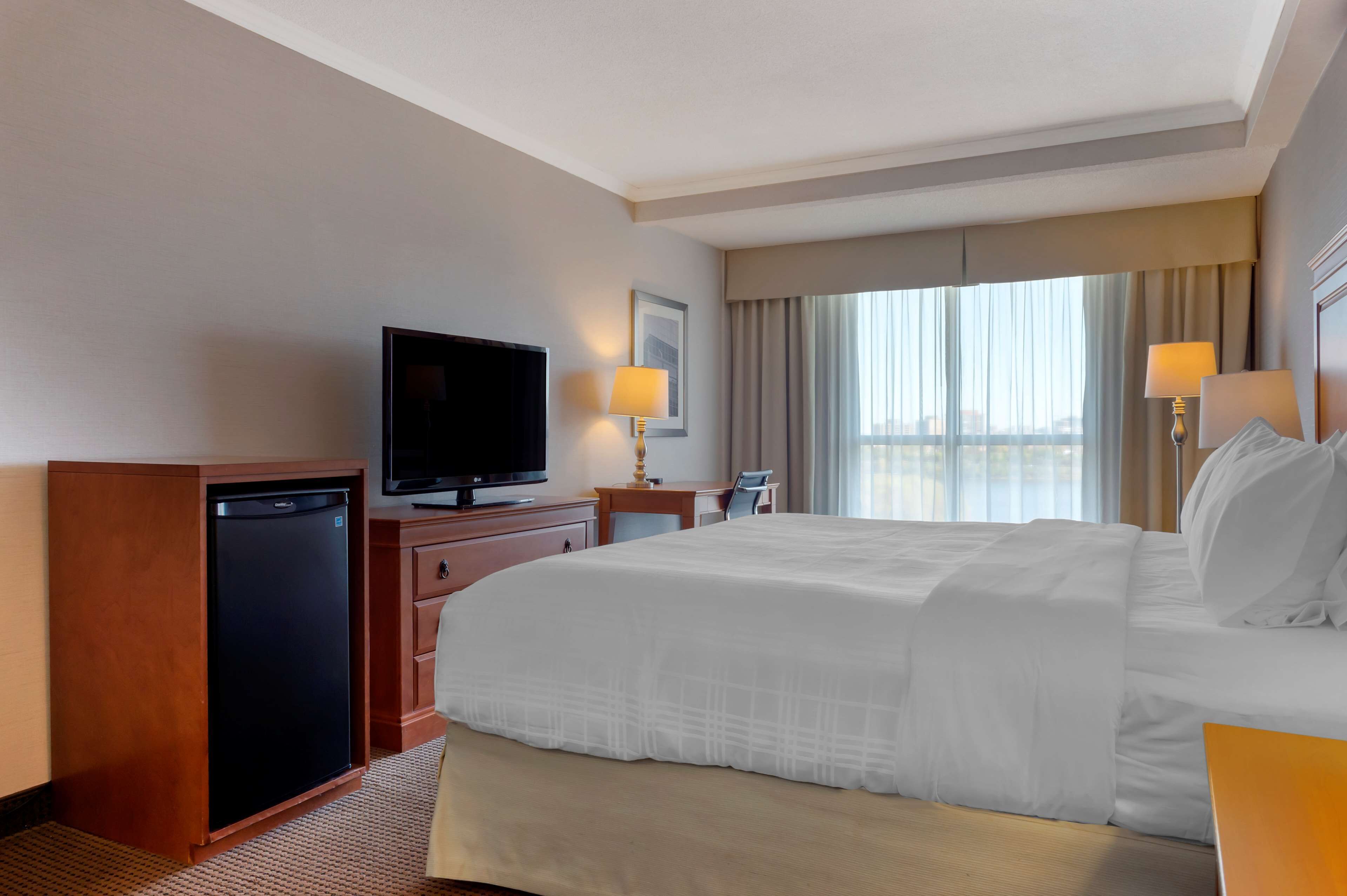 A hotel room with a large bed, a television, a desk with a lamp, and a view of the city through the window.