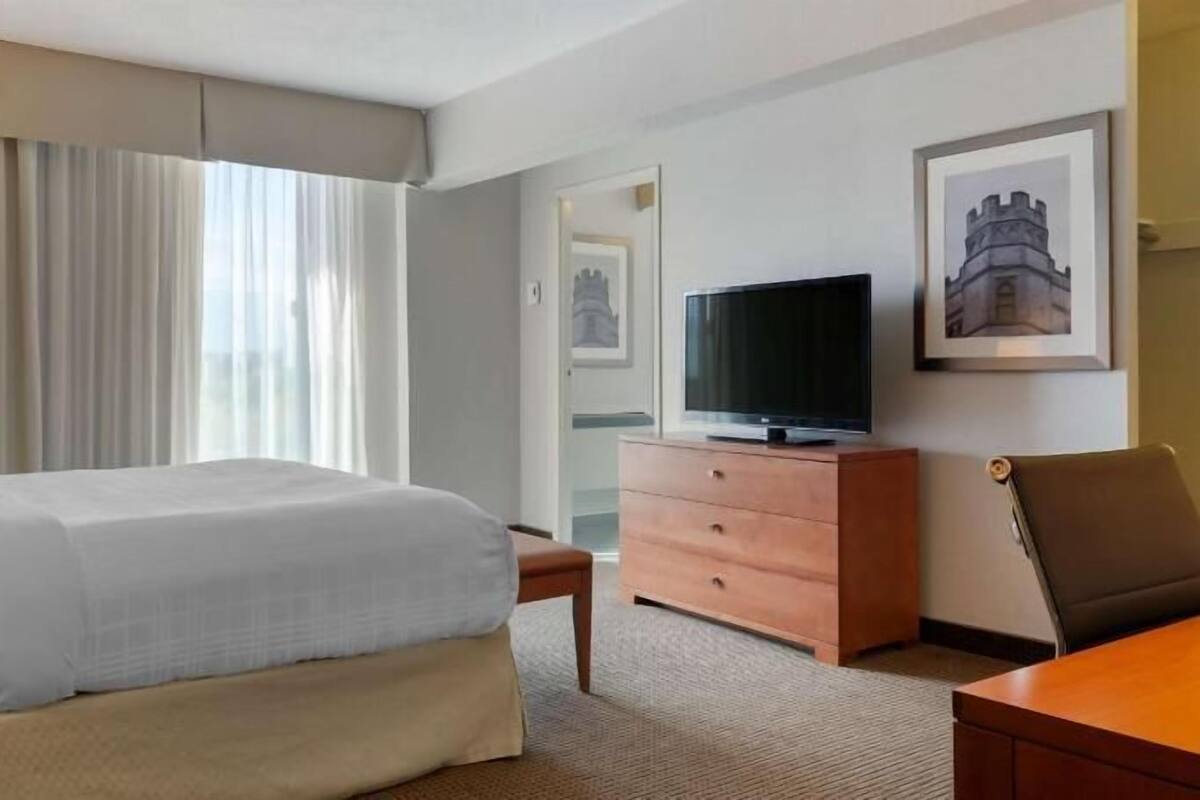 Ein Hotelzimmer mit einem Bett, einem Fernseher, einem Schminktisch, einem Stuhl und einem gerahmten Bild an der Wand.