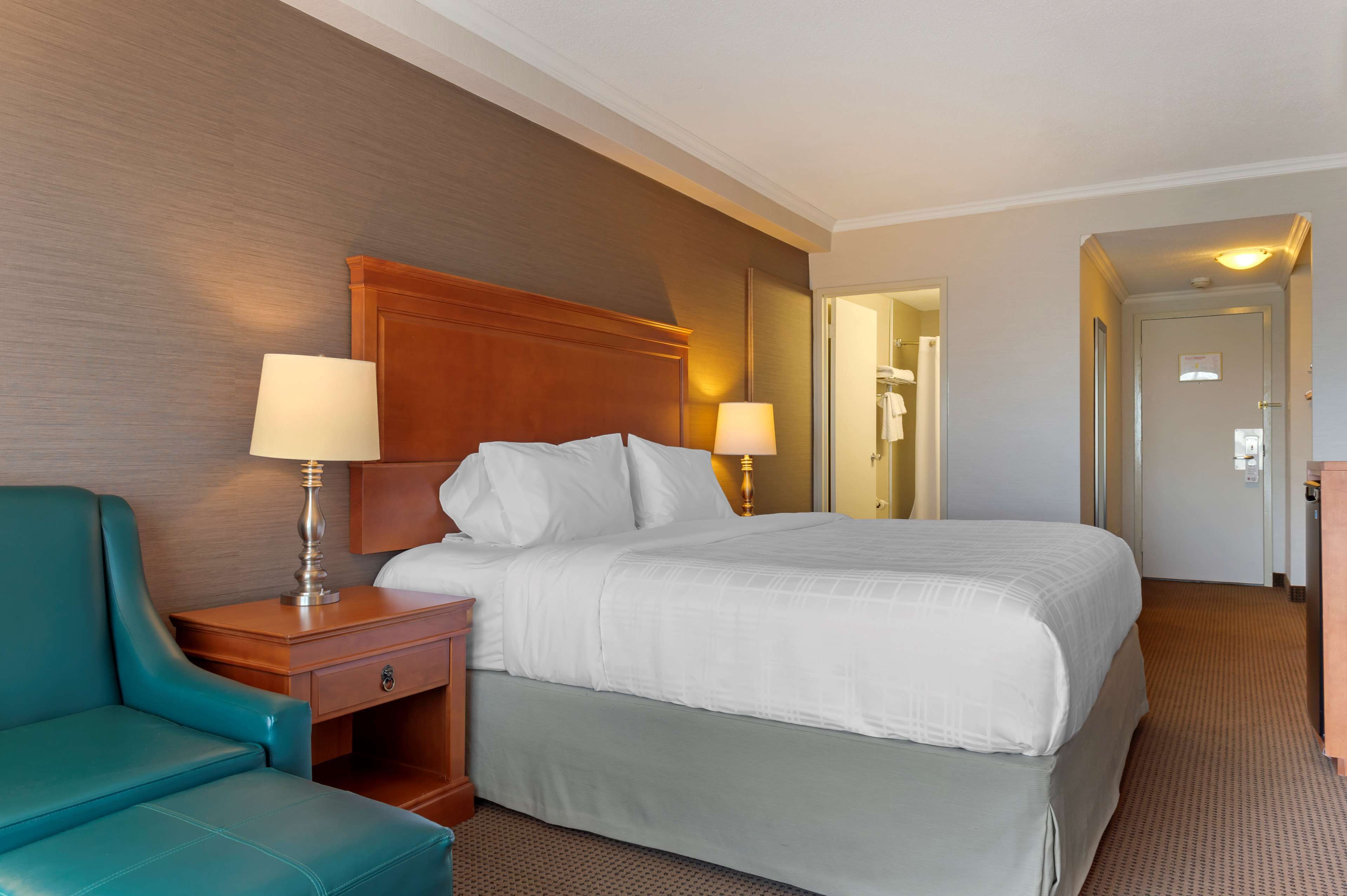 A hotel room with a large bed, two bedside lamps, a wooden headboard, a teal armchair, and a mirror on the wall.