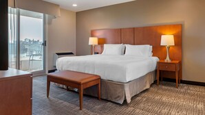A hotel room with a large bed, two bedside lamps, a wooden bench, a desk, and a large window offering a city view.
