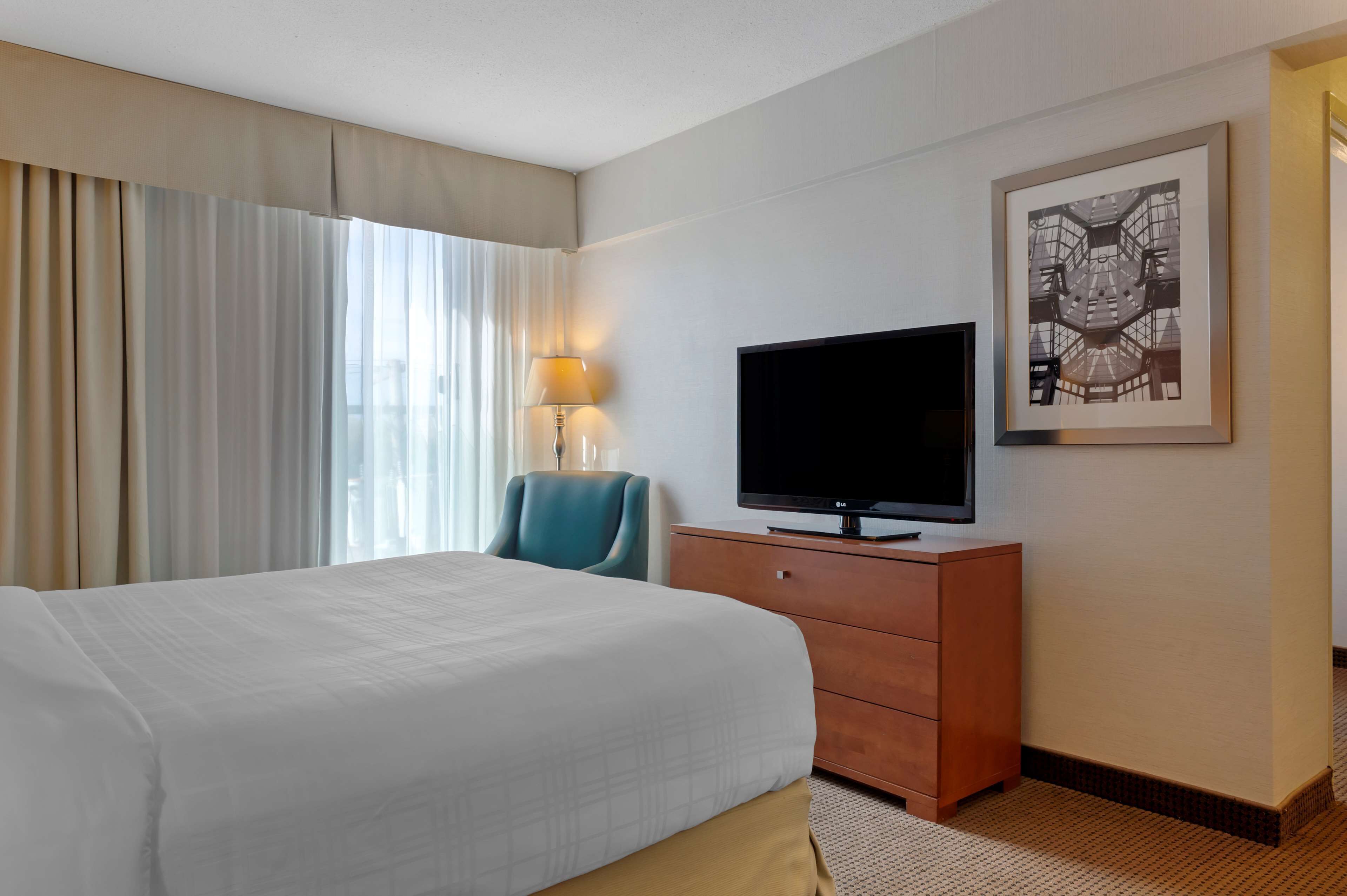 Une chambre d’hôtel avec un grand lit, un téléviseur posé sur une commode en bois, une lampe et un tableau encadré au mur.