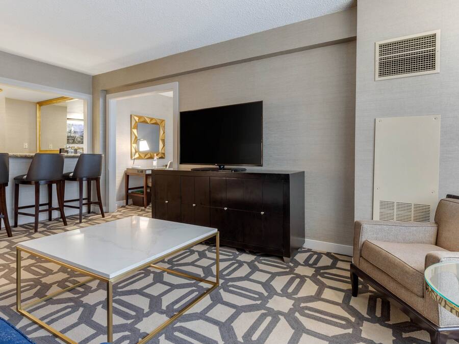 A modern hotel room with a dining area, a flat-screen TV, a sofa, and a patterned carpet.